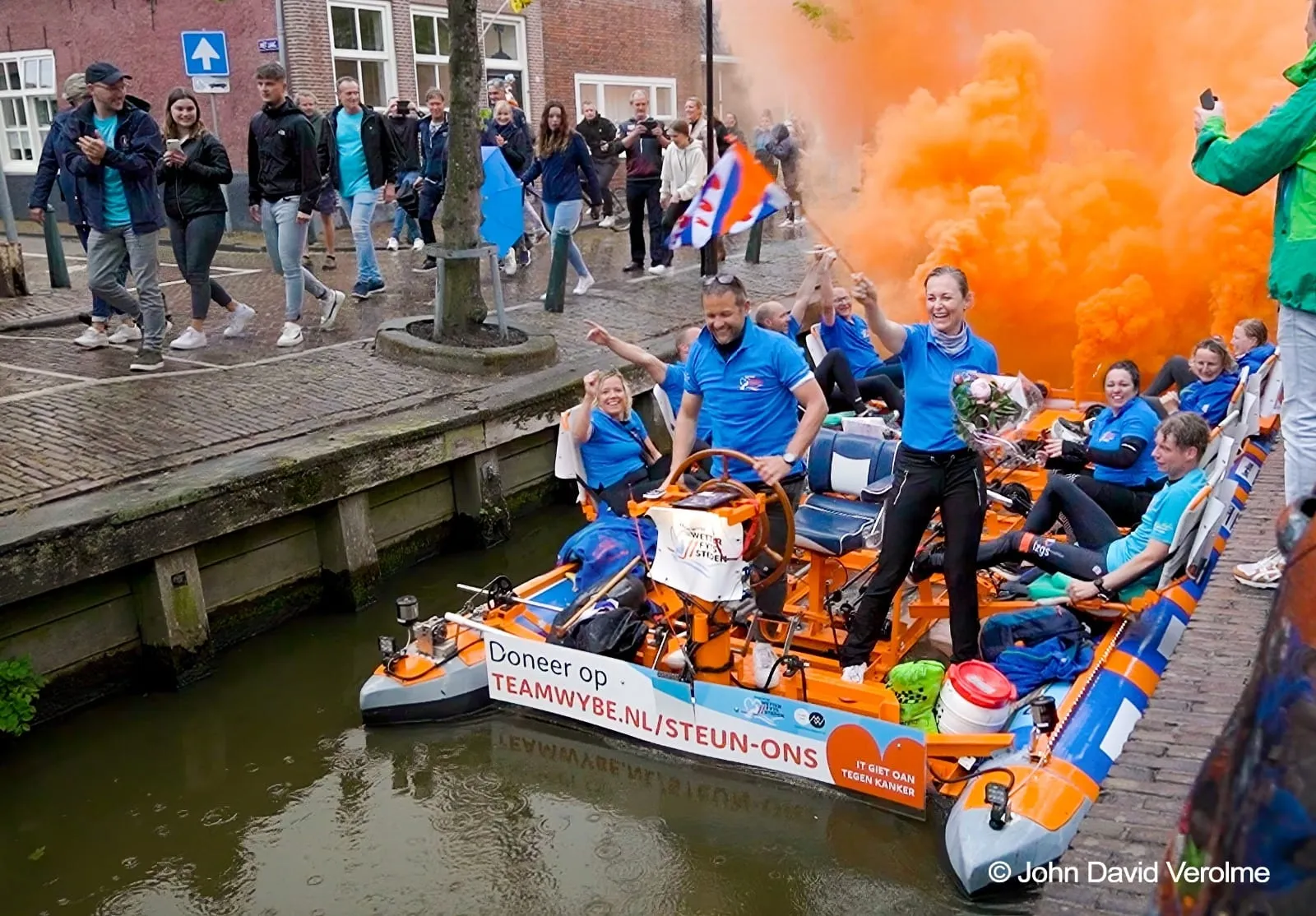 De wetterfyts van Team Wybe vaart onder oranje rook door een Friese stad in 2022